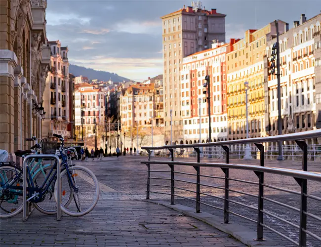 Avenida em cidade europeia. Há prédios diversos e uma estação para bicicletas, alternativa sustentável de transporte.